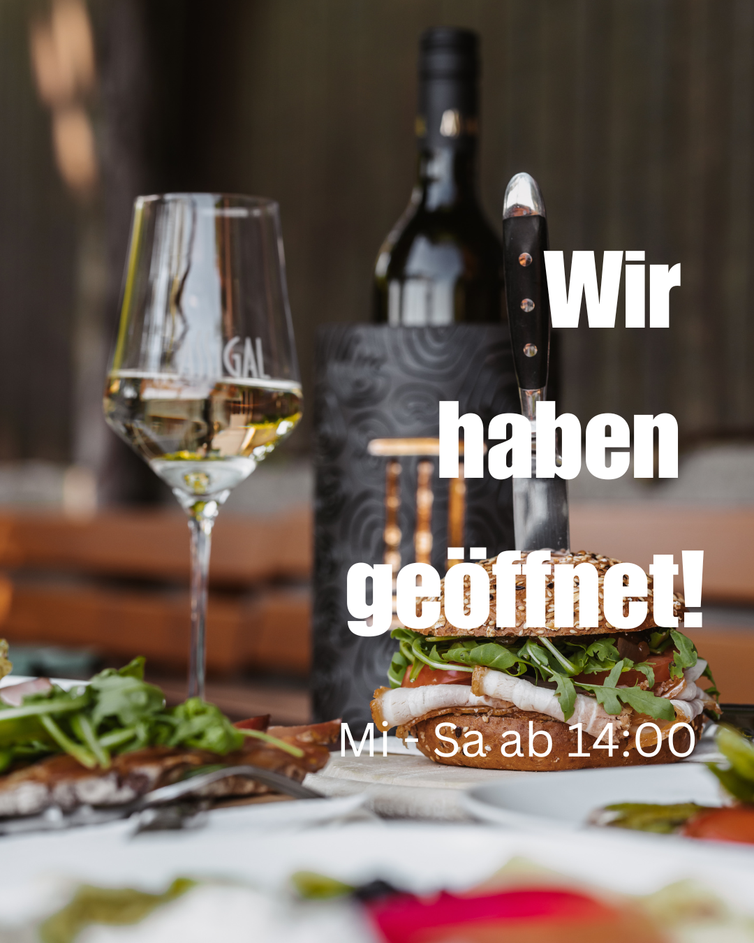Eine Flasche Wein, ein Messer, ein Glas Wein und ein Sandwich mit Gemüse stehen auf dem Tisch. Der Text lautet 'Wir haben geöffnet! Ab Samstag um 14:00 Uhr.'