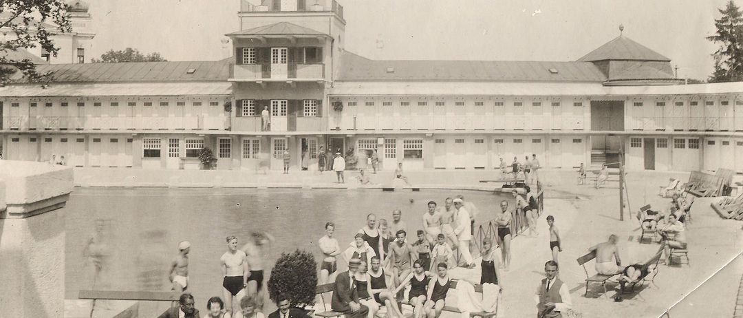 Ein Schwarz-Weiß-Foto zeigt Menschen an einem Schwimmbad. Ein Uhrturm steht über dem Pool mit einem Gebäude dahinter.