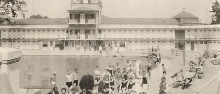 Ein Schwarz-Weiß-Foto zeigt Menschen an einem Schwimmbad. Ein Uhrturm steht über dem Pool mit einem Gebäude dahinter.