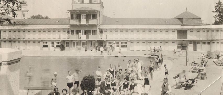 Ein Schwarz-Weiß-Foto zeigt Menschen an einem Schwimmbad. Ein Uhrturm steht über dem Pool mit einem Gebäude dahinter.
