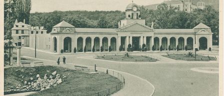 Eine alte Postkarte zeigt ein großes Gebäude namens Grand Hotel. Es hat viele Säulen, einen Uhrturm und ist von Bäumen umgeben. Zwei Personen gehen davor.