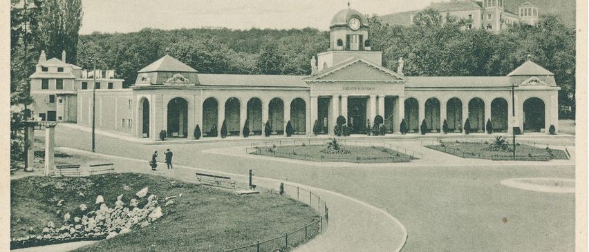Eine alte Postkarte zeigt ein großes Gebäude namens Grand Hotel. Es hat viele Säulen, einen Uhrturm und ist von Bäumen umgeben. Zwei Personen gehen davor.
