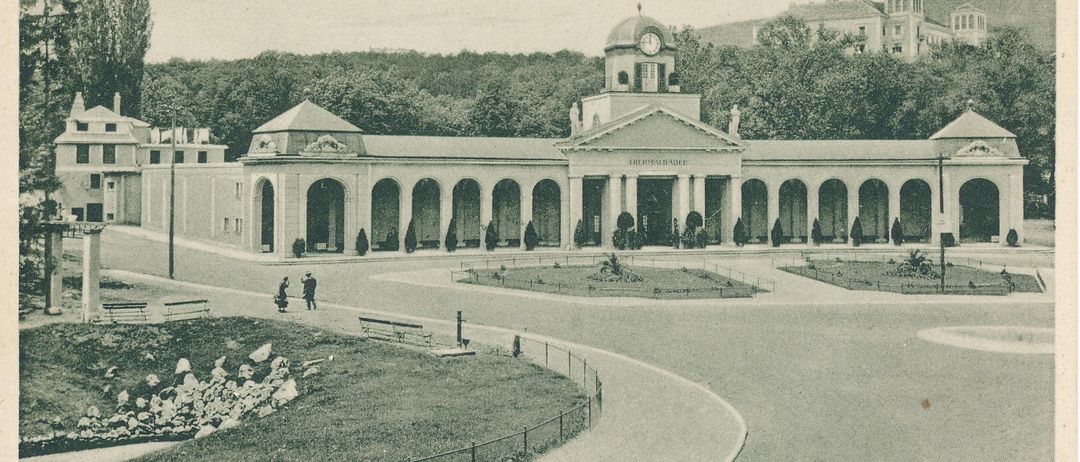 Eine alte Postkarte zeigt ein großes Gebäude namens Grand Hotel. Es hat viele Säulen, einen Uhrturm und ist von Bäumen umgeben. Zwei Personen gehen davor.