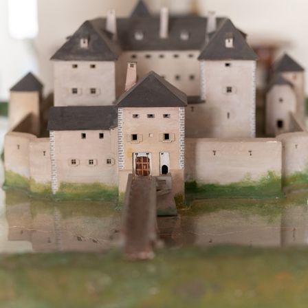 Ein Miniaturmodell einer mittelalterlichen Burg mit Türmen und einer Zugbrücke, auf einer reflektierenden Oberfläche mit einem grasbewachsenen Bereich darum platziert.