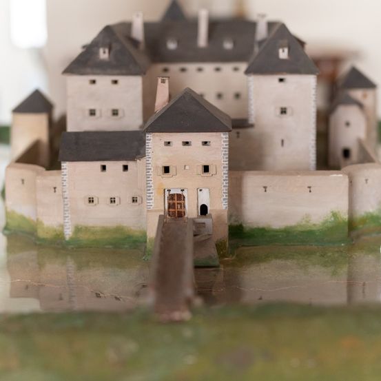 Ein Miniaturmodell einer mittelalterlichen Burg mit Türmen und einer Zugbrücke, auf einer reflektierenden Oberfläche mit einem grasbewachsenen Bereich darum platziert.