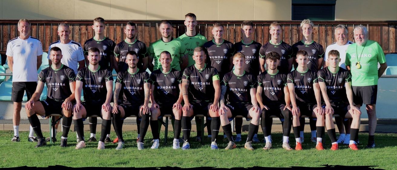 Das Fußballteam posiert für ein Foto vor dem 'Fans Tropper Stadion' mit den Jahren 2025/26 und dem Sponsorennamen 'Wundara' sichtbar.