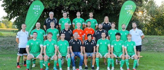 Eine Fußballmannschaft in grünen Trikots posiert für ein Gruppenfoto auf einem Rasenfeld mit Bannern dahinter.