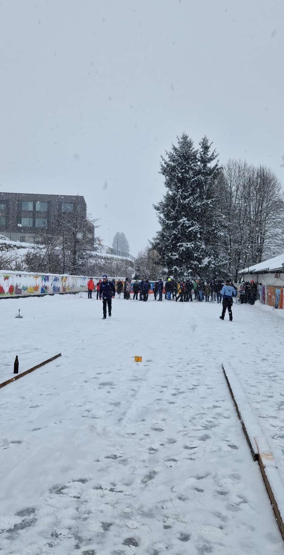 In einem verschneiten Außenbereich versammeln sich mehrere Menschen um eine Curlingbahn. Ein Gebäude befindet sich hinter ihnen, und Bäume umgeben das Gebiet. Der Boden ist mit Schnee bedeckt.