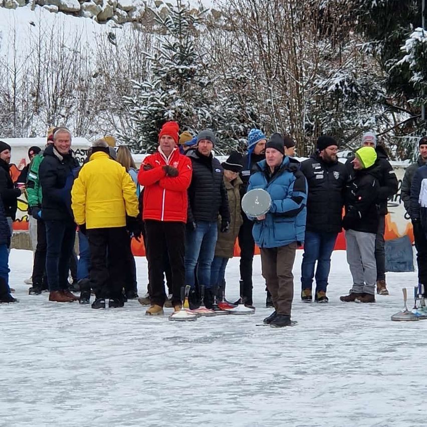 Eine Gruppe von Menschen steht auf einem verschneiten Feld, einige tragen Winterkleidung, Mützen und Handschuhe. Ein Mann hält einen Scheiben. Hinter ihnen sind schneebedeckte Bäume und eine weiße Barriere.