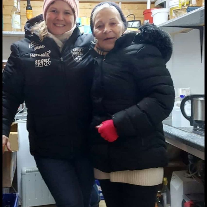 Zwei Frauen stehen eng beieinander in einer Küche, beide tragen schwarze Jacken. Die Frau auf der linken Seite lächelt und trägt eine Mütze. Die Frau auf der rechten Seite trägt rote Handschuhe. Im Hintergrund stehen Regale mit Flaschen und Behältern.