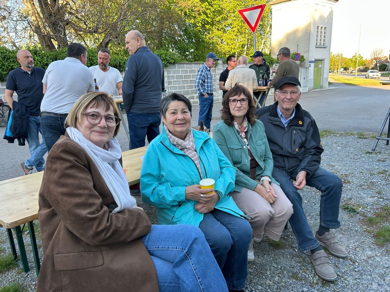 Eine Gruppe von Menschen sitzt draußen auf einer Bank. Einige tragen Brillen, Jacken und Schals. Eine Frau hält einen Becher. Hinter ihnen stehen oder sitzen andere, und ein Gebäude steht im Hintergrund.
