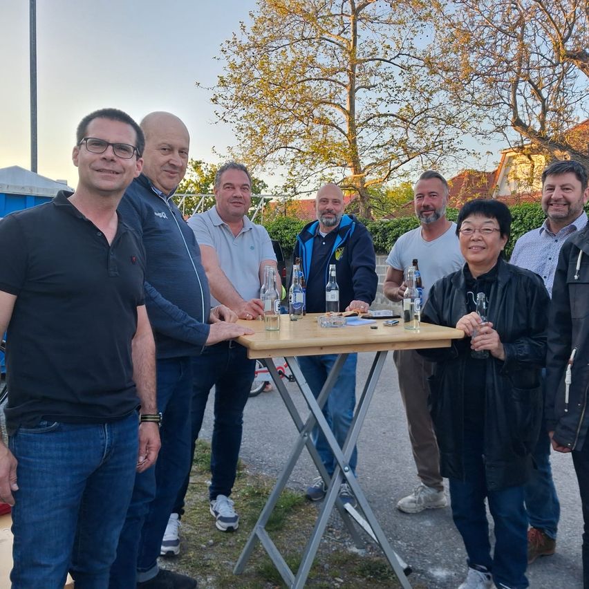 Eine Gruppe von Männern und einer Frau steht lächelnd um einen Tisch mit Flaschen. Sie befinden sich in einer Außenumgebung mit Bäumen und Gebäuden im Hintergrund.