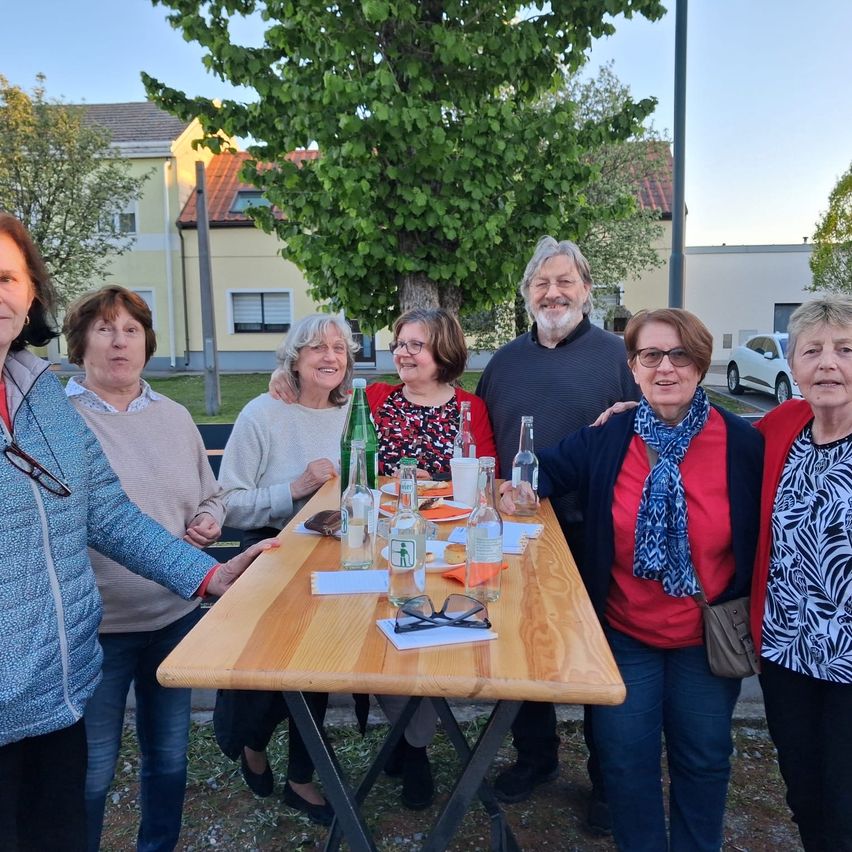 Eine Gruppe Erwachsener, darunter Frauen und Männer, steht im Freien um einen Tisch mit verschiedenen Gegenständen wie Gläsern, Flaschen und Servietten.