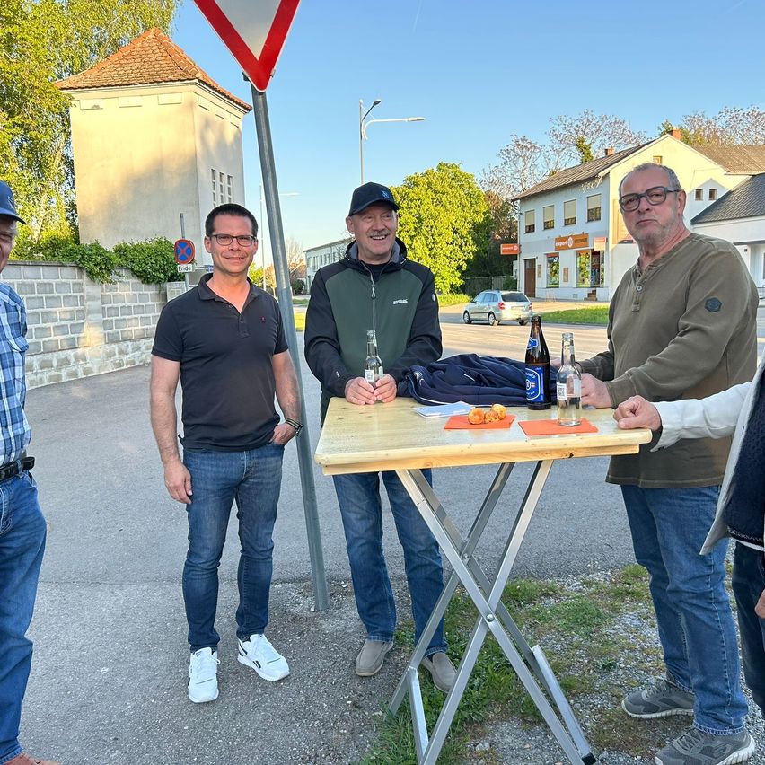 Fünf Männer stehen um einen kleinen Holztisch auf einer Straße, halten Getränke und lächeln. Dahinter steht ein Gebäude mit Bäumen und einem Straßenschild.