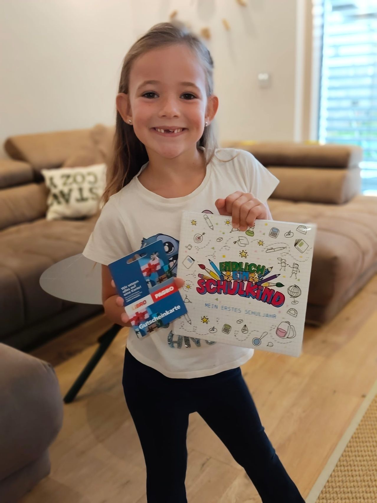 Ein kleines Mädchen hält ein Buch und eine Geschenkkarte in der Hand und lächelt in einem gemütlichen Wohnzimmer.