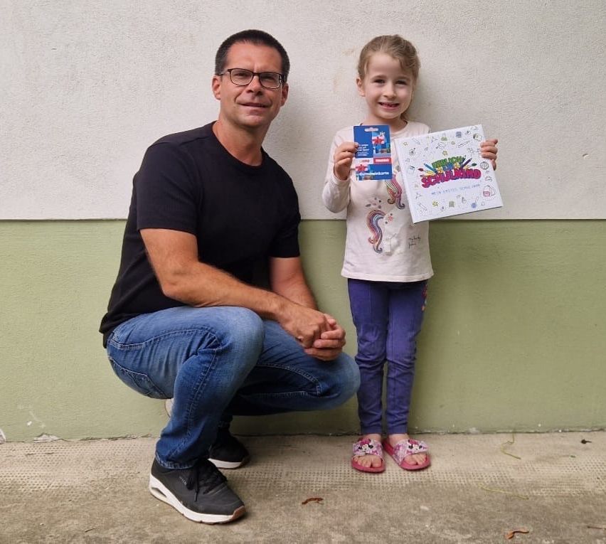 Ein Mann und ein kleines Mädchen posieren für ein Foto, während sie Bücher halten. Der Mann kniet auf dem Boden und das Mädchen steht neben ihm. Sie lächeln beide.