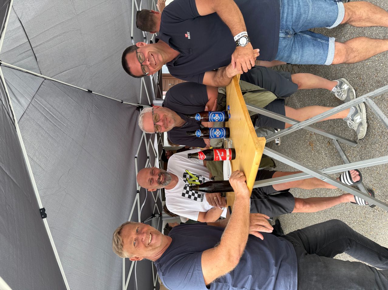Fünf Männer lächeln und posieren für ein Foto bei einer Outdoor-Veranstaltung. Flaschen Bier stehen auf einem Tisch vor ihnen.