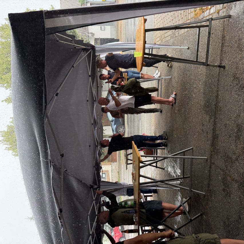 Eine Gruppe von Menschen sitzt unter einem großen Zelt auf einem nassen Bürgersteig, mit mehreren Tischen und Stühlen in einem Outdoor-Essbereich aufgestellt.