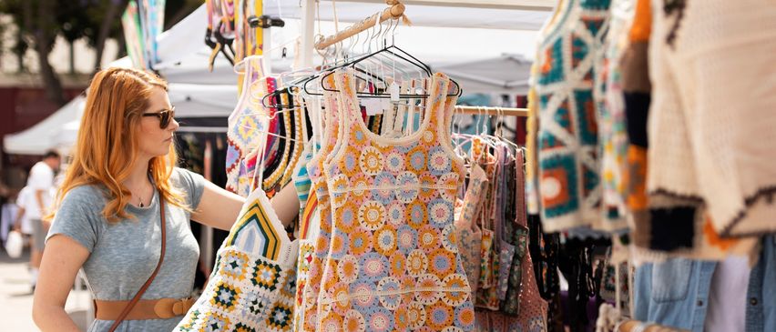 Eine Frau in einem grauen Kleid stöbert in einem Stand voller bunter gehäkelte Kleider und Taschen auf einem Markt.