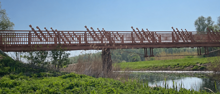 Eine rote Holzbrücke überspannt einen ruhigen Wasserlauf, umgeben von saftigem grünem Gras und Bäumen unter einem klaren blauen Himmel.