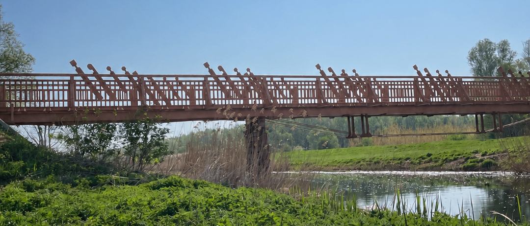 Eine rote Holzbrücke überspannt einen ruhigen Wasserlauf, umgeben von saftigem grünem Gras und Bäumen unter einem klaren blauen Himmel.