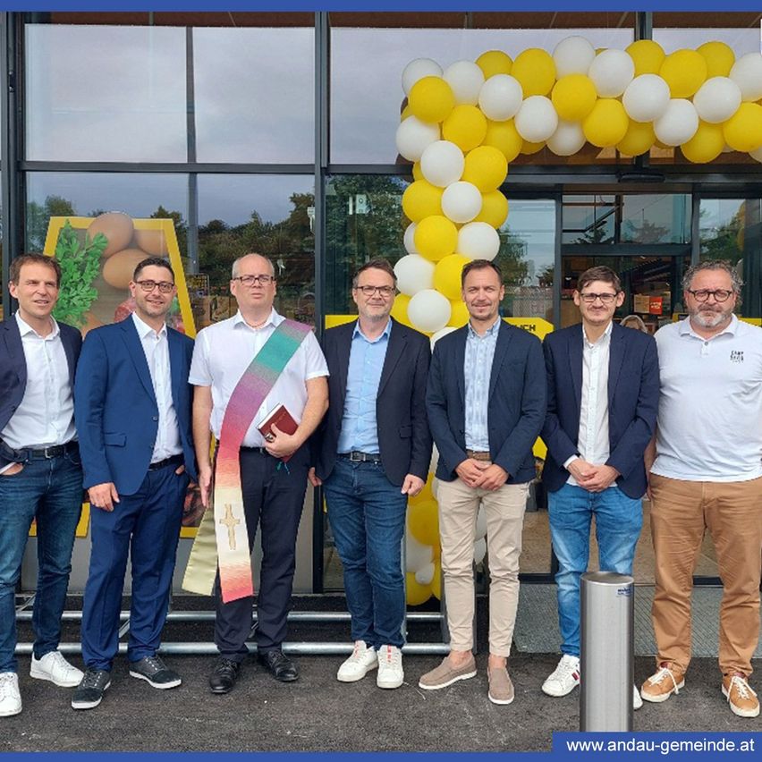 Eine Gruppe von Männern in Geschäftskleidung steht vor dem Eingang eines Geschäfts mit einem gelben und weißen Luftballonbogen. Ein Mann hält ein Buch und eine Schärpe.