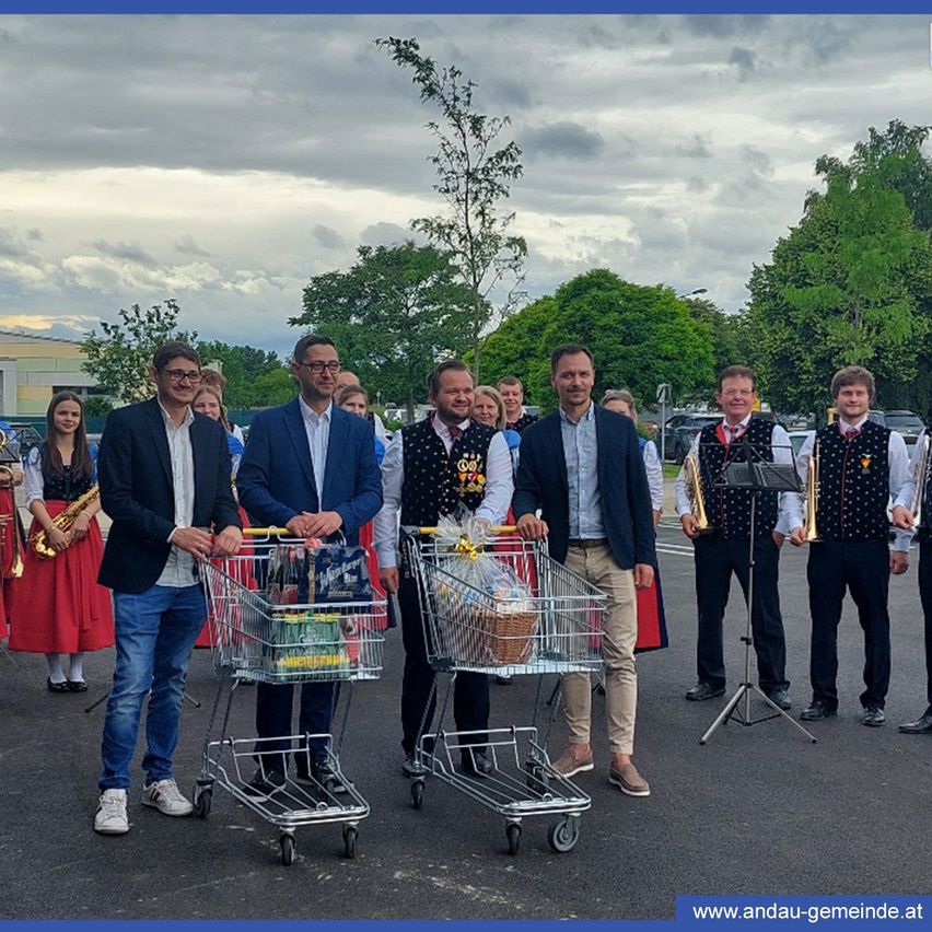 Eine Gruppe von Menschen mit Einkaufswagen auf einem Parkplatz. Hinter ihnen stehen Musiker mit Instrumenten. Eine Person spielt die Saxophon. Im Hintergrund befinden sich Bäume und ein bewölkter Himmel.