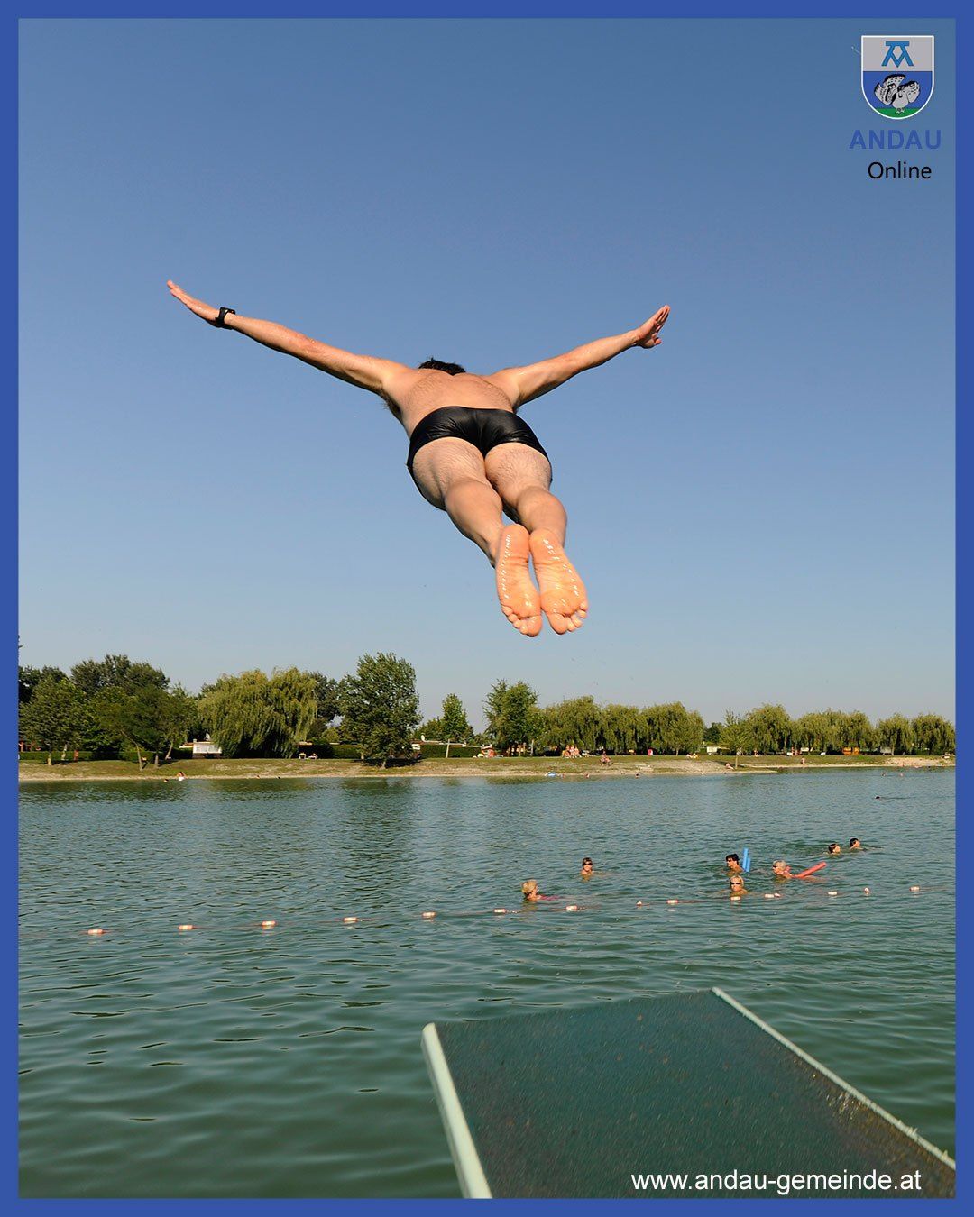 Ein Mann springt in einen See, während mehrere Menschen in seiner Nähe schwimmen, unter einem klaren blauen Himmel.