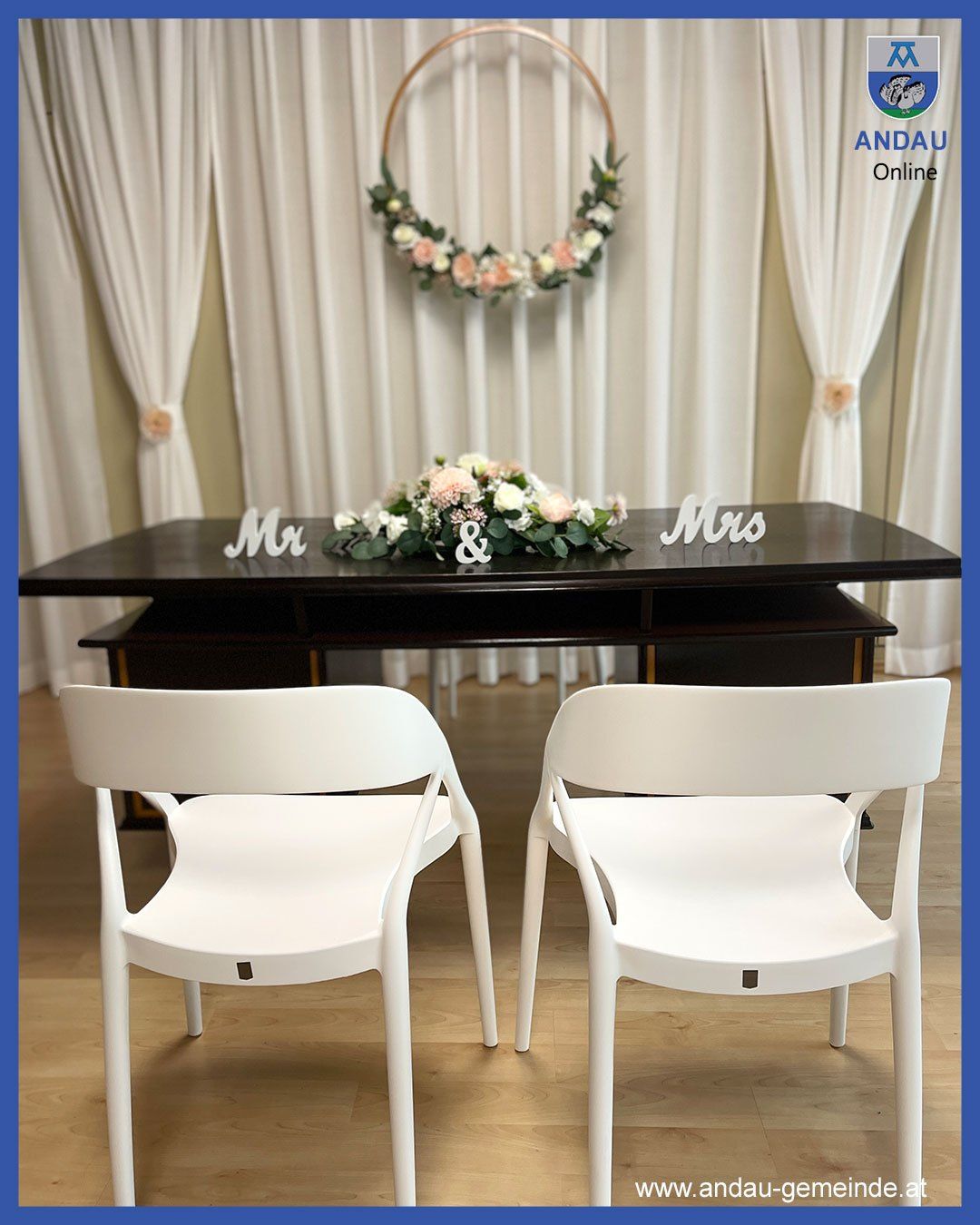 Eine Hochzeitsdekoration mit einem Tisch und zwei Stühlen, geschmückt mit einem Blumenarrangement und Mr. und Mrs. Schildern, vor einem weißen Vorhang als Hintergrund.