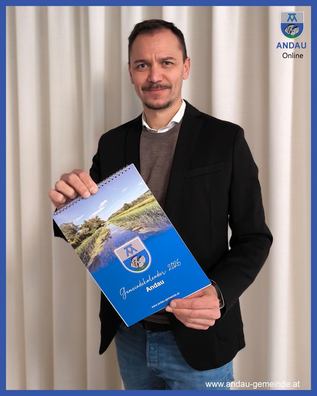 Ein Mann in einem schwarzen Anzug hält ein blaues Spiralnotizbuch mit der Titelseite eines Sees und eines Logos, betitelt 'Gemeindetalenten 2016 Andau'. Er lächelt und scheint für ein Foto zu posieren.