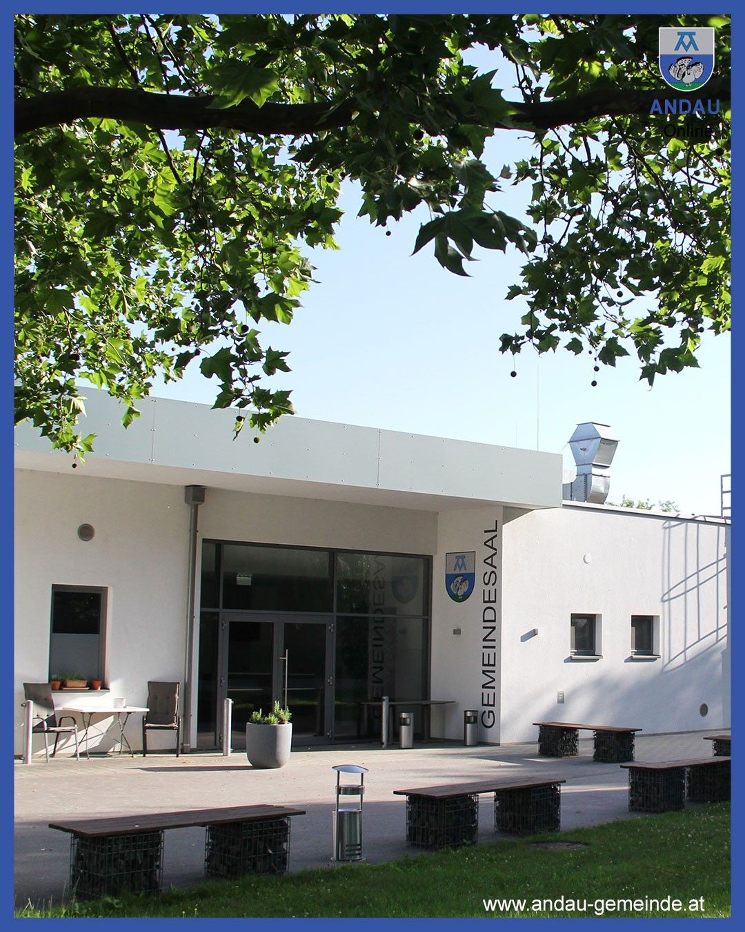 Die Vorderseite eines weißen Gebäudes mit einem Schild, das Gemeindesaal steht. Draußen sind Bänke, ein Tisch und Stühle. Ein Topf mit Pflanzen und Mülleimer stehen vor dem Eingang.