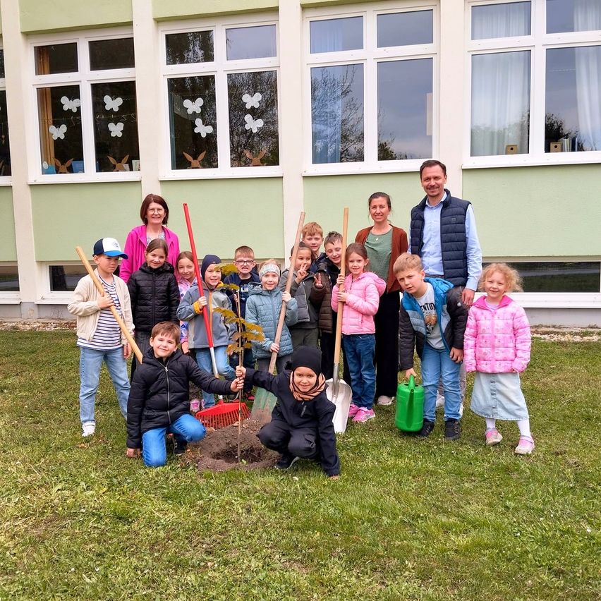 Eine Gruppe von Kindern und Erwachsenen posiert vor einem Schulgebäude, wobei sie im Vordergrund einen Baum pflanzen. Die Erwachsenen halten Schaufeln und Rechen, und einige Kinder tragen Mützen.