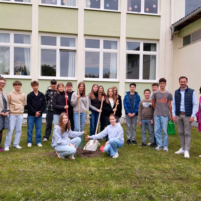 Eine Gruppe von Menschen, darunter Schüler, pflanzt einen Baum vor einem Gebäude. Sie tragen alle Freizeitkleidung und lächeln.