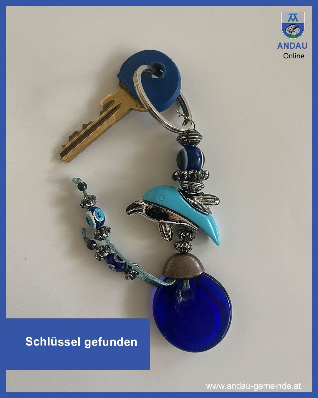 Ein blauer Schlüsselanhänger mit einem silbernen Delfin-Anhänger hängt an einem goldenen Schlüssel auf einer weißen Oberfläche. Unter dem Schlüsselanhänger wird der Text 'Schlüssel gefunden' angezeigt.