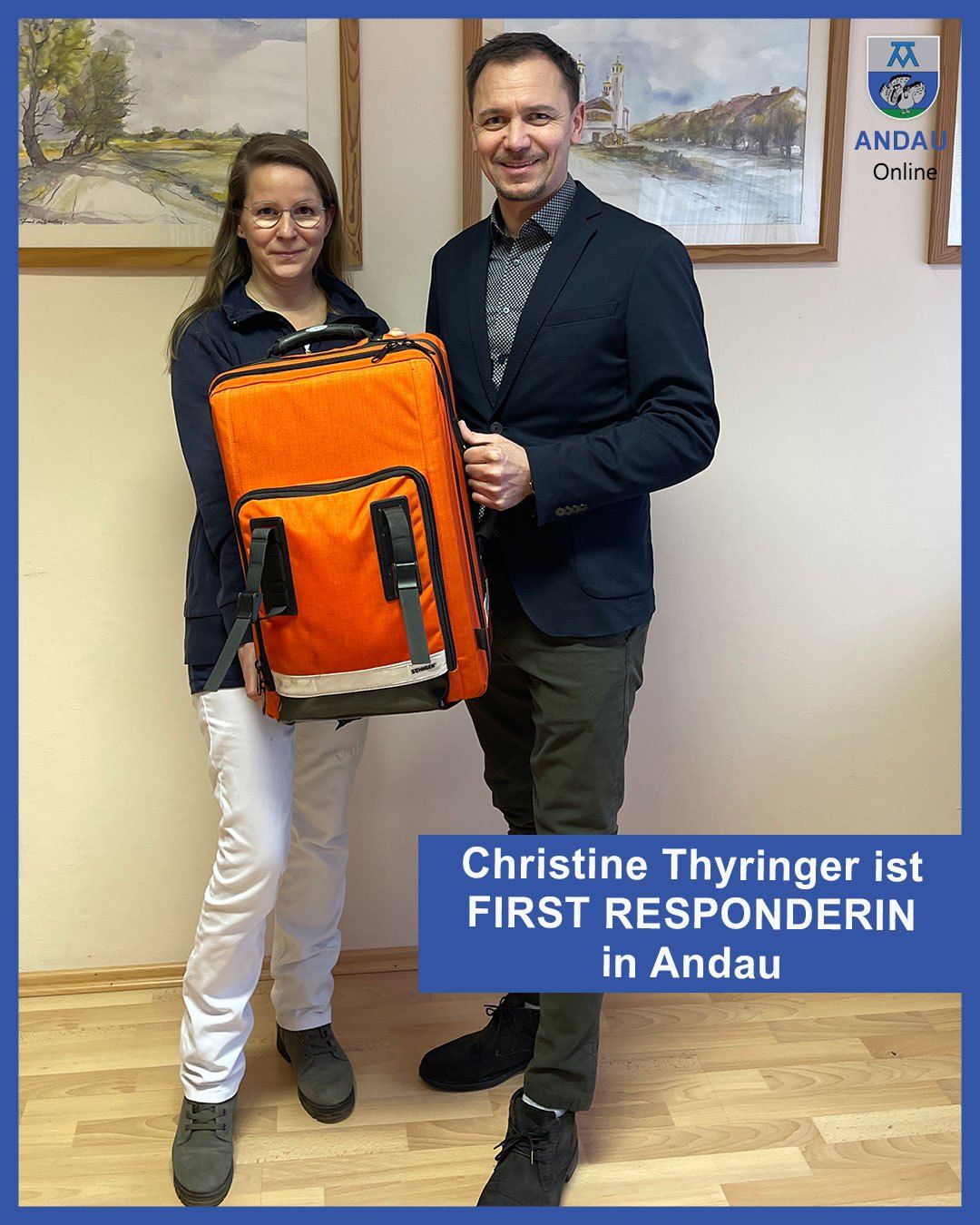 Christine Thyringer, wearing glasses and a jacket, is holding an orange bag with a man in a suit and tie. They are standing in front of a white wall. Christine is the first responder in Andau.