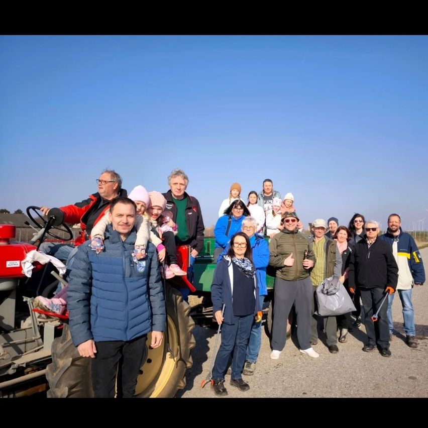 Eine Gruppe von Menschen steht vor einem Traktor und posiert fr ein Foto. Einige tragen Brillen, Hte und Tcher. Ein Mann vorne hlt ein Werkzeug.