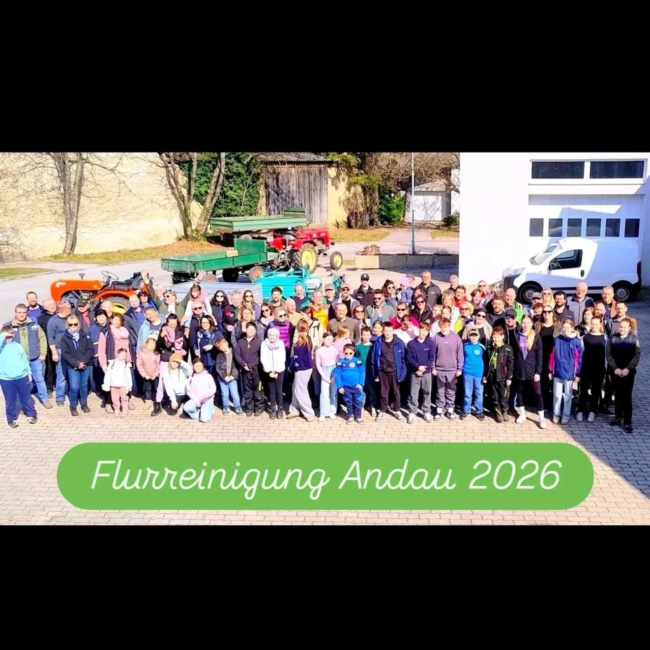 Eine große Gruppe von Menschen posiert für ein Foto vor einem Gebäude. Traktoren und ein Lieferwagen sind dahinter geparkt.