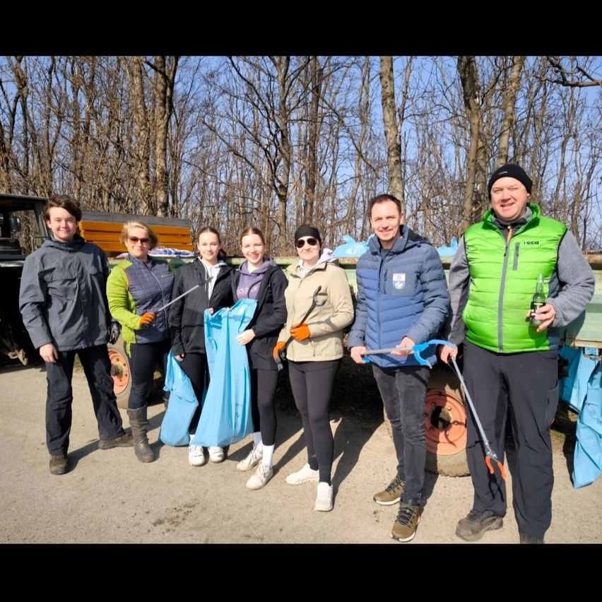 Eine Gruppe von Menschen in warmer Kleidung, die in einem Wald stehen und Werkzeuge und Müllsäcke halten. Ein Mann hält ein Bier. Sie lächeln für ein Foto.
