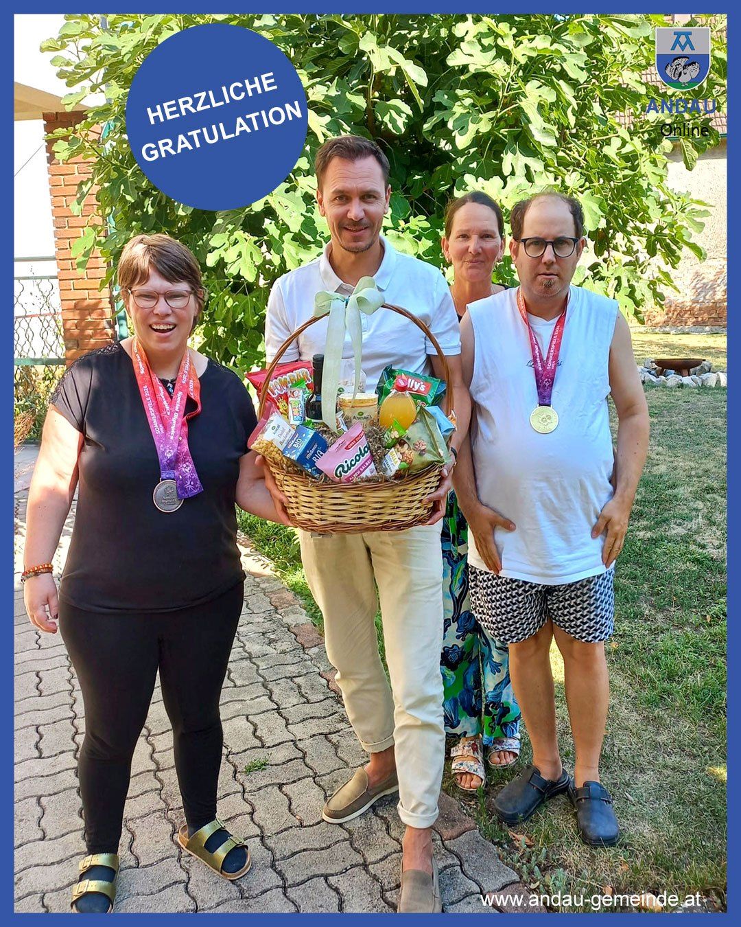 Eine Gruppe von vier Personen steht draußen, einer hält einen Korb mit Snacks, alle tragen Medaillen. Der Mann in der Mitte lächelt und die Frau links lacht. Hinter ihnen steht eine Ziegelmauer und ein Baum.