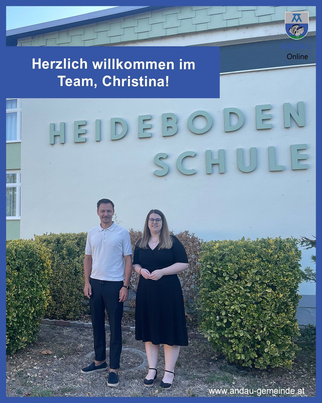 Ein Mann und eine Frau stehen vor einem Gebäude mit dem Schild 'HEIDEBODEN SCHUL'. Der Mann trägt ein weißes Poloshirt und schwarze Hose, die Frau ein schwarzes Kleid. Sie lächeln für ein Foto.