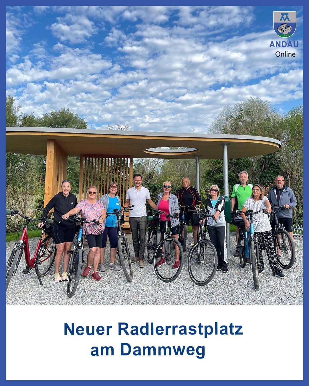 Eine Gruppe von Menschen in Fahrradausrüstung posiert für ein Foto vor einem neuen Fahrradrastplatz. Der Bereich befindet sich unter einem modernen Vordach mit einem klaren blauen Himmel im Hintergrund.