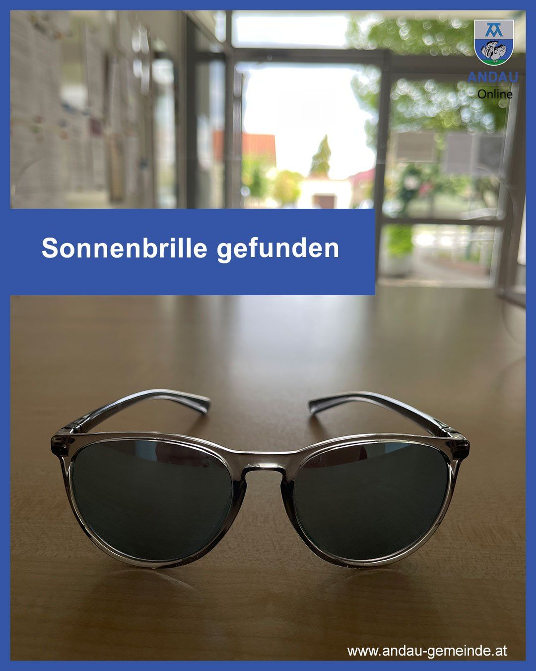 Ein Paar Sonnenbrille mit schwarzem Rahmen und reflektierenden Gläsern liegt auf einem Holztisch. Hinter der Sonnenbrille befindet sich ein blauer Banner mit weißem Text Sonnenbrille gefunden. Der Hintergrund ist unscharf.