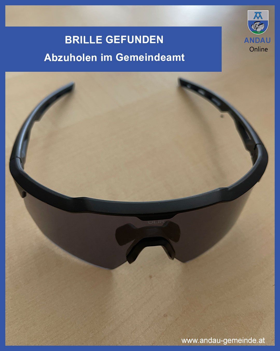 A pair of black sunglasses with a black frame and dark lenses lies on a brown surface. A blue banner with white text reads Brille Gefunden and Abzuholen im Gemeindeamt.
