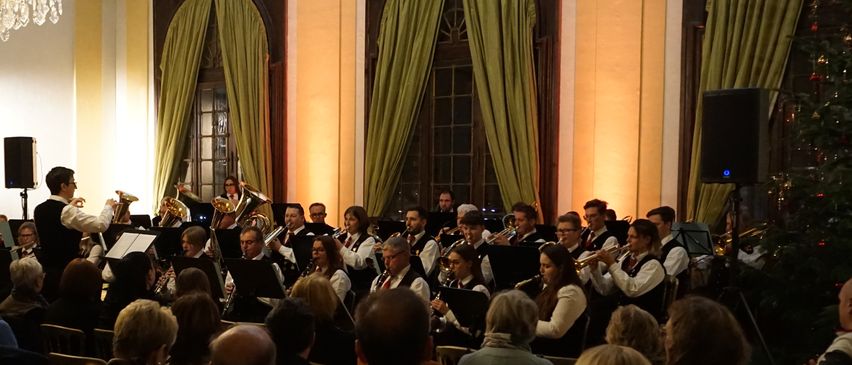 Ein Konzert findet in einem Saal mit Kronleuchtern statt. Die Musiker spielen vor einem Publikum, das auf Stühlen sitzt.