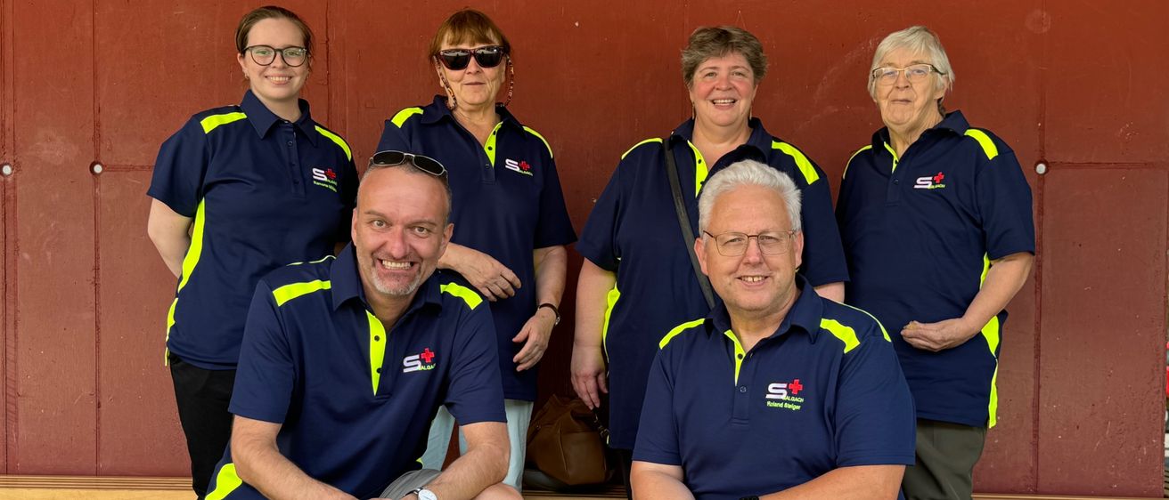 A group of six individuals in matching blue and yellow uniforms with an 'S+' logo on the left chest, posing for a photo in front of a red wall, with two men kneeling and four women standing behind them.