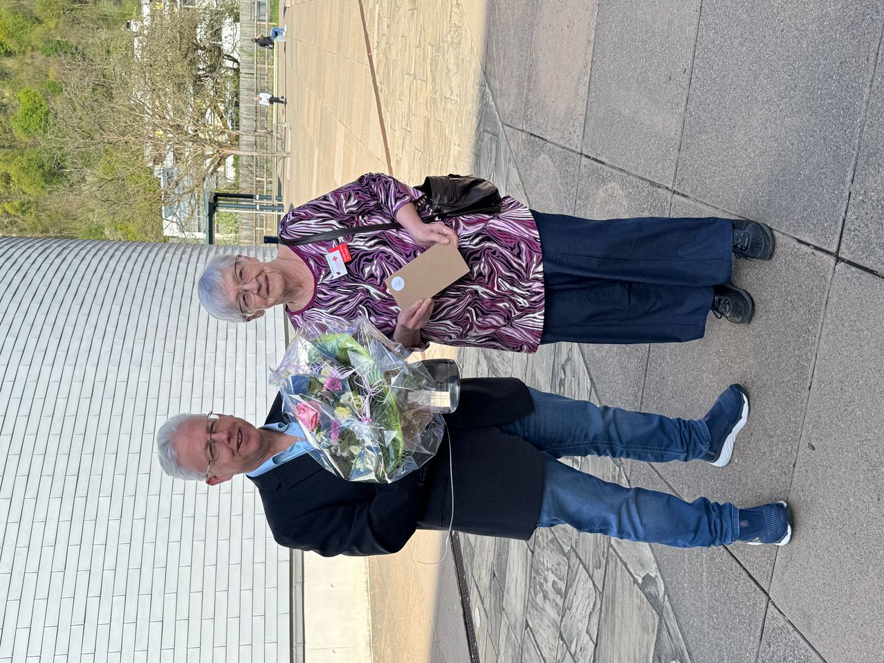 Two individuals stand on a concrete pathway, holding a bouquet of flowers. An elderly woman, wearing glasses and a purple shirt, smiles. The man beneath her holds the bouquet, and both are dressed casually. Trees and buildings are visible in the background.