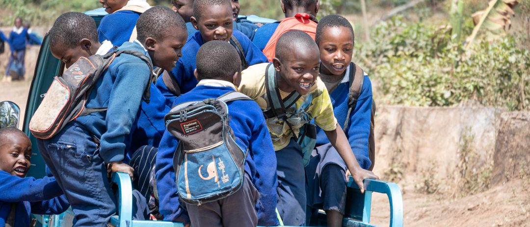 Eine Gruppe von Kindern in blauen Uniformen fährt im hinteren Teil eines blauen Trucks im Freien. Sie lächeln und scheinen sich zu amüsieren.