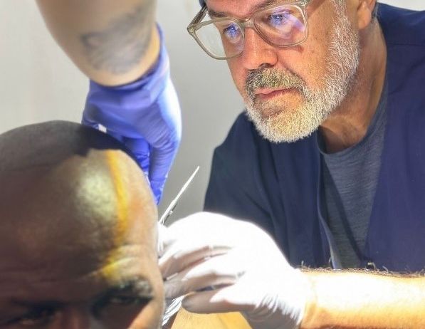 Ein Mann mit kahlem Kopf und gelben Markierungen auf der Stirn bekommt einen Haarschnitt von einer Person in medizinischer Kleidung, Handschuhen und Brille.