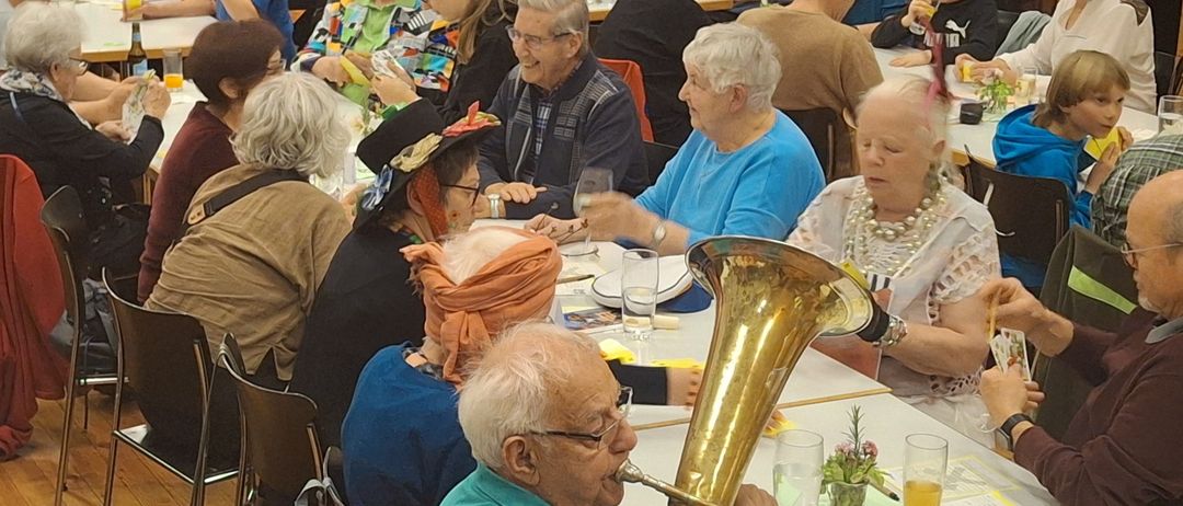 Eine Gruppe älterer Menschen trifft sich zu einem Essensevent, einige tragen Hüte und Brillen. Ein älterer Mann spielt ein Blechinstrument, während andere ihre Mahlzeiten genießen. Der Raum hat Holzböden und große Fenster mit hängenden Dekorationen.
