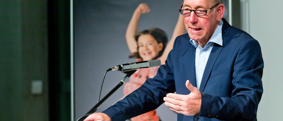 Ein Mann steht an einem Podium und spricht in ein Mikrofon mit einem Schild, das '4' vor ihm zeigt, mit einem Bild eines Kindes im Hintergrund.